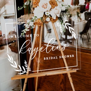 May include: A clear acrylic sign with white calligraphy that reads "Welcome to Kayelin's Bridal Shower" on a wooden easel with a bouquet of dried flowers in front of it.