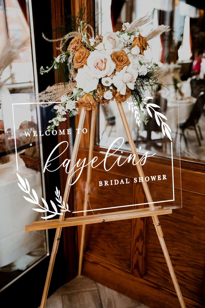 May include: A clear acrylic sign with white calligraphy that reads "Welcome to Kayelin's Bridal Shower" on a wooden easel with a bouquet of dried flowers in front of it.