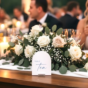 May include: A wedding reception table setting with floral arrangements of white roses, greenery, and candles. A sign that reads "Table Ten" is centered on the table. Guests are seated in the background.