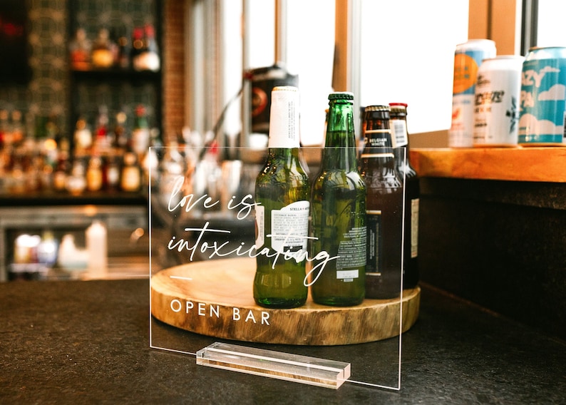 May include: A clear acrylic sign with the text "Love is intoxicating" and "Open Bar" on a wooden base. The sign is in front of a bar with bottles of alcohol and cans of beer.