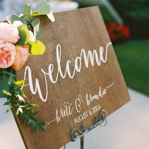 May include: A wooden welcome sign with white calligraphy that reads "welcome" and "brett & brandie" with the date "august 6, 2016". The sign is decorated with greenery and pink flowers.