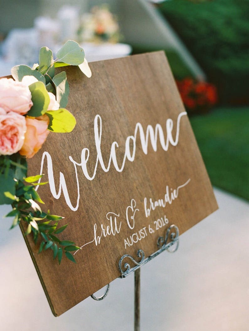 May include: A wooden welcome sign with white calligraphy that reads "welcome" and "brett & brandie" with the date "august 6, 2016". The sign is decorated with greenery and pink flowers.