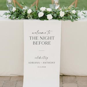 May include: A wedding reception table is decorated with a floral arrangement of white roses and greenery. A white banner reads "welcome to THE NIGHT BEFORE celebrating ADRIANA + ANTHONY 3.4.2024". Two wooden chairs and glasses are in the background.