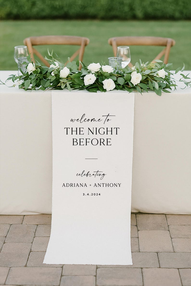 May include: A wedding reception table is decorated with a floral arrangement of white roses and greenery. A white banner reads "welcome to THE NIGHT BEFORE celebrating ADRIANA + ANTHONY 3.4.2024". Two wooden chairs and glasses are in the background.