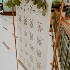 May include: A white sign with black text on a copper stand with greenery. The sign says "Please Find Your Seat" and lists names for each table number.
