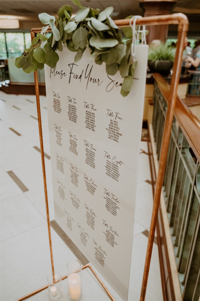 May include: A white sign with black text on a copper stand with greenery. The sign says "Please Find Your Seat" and lists names for each table number.