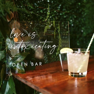 May include: A clear acrylic sign with the text "Love is intoxicating" and "Open Bar" written in white calligraphy. A glass of lemonade with a lime wedge and a green and white striped straw sits on a wooden table.