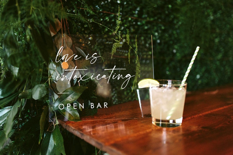 May include: A clear acrylic sign with the text "Love is intoxicating" and "Open Bar" written in white calligraphy. A glass of lemonade with a lime wedge and a green and white striped straw sits on a wooden table.