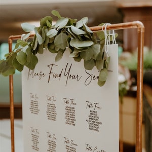 May include: A white sign with black text listing table numbers and guest names for a wedding reception. The sign is hanging from a copper pipe stand and decorated with greenery.