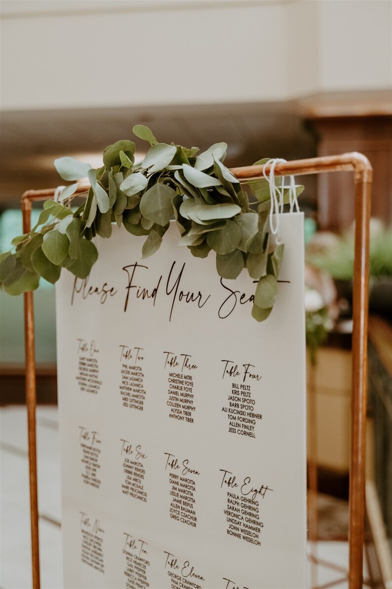May include: A white sign with black text listing table numbers and guest names for a wedding reception. The sign is hanging from a copper pipe stand and decorated with greenery.