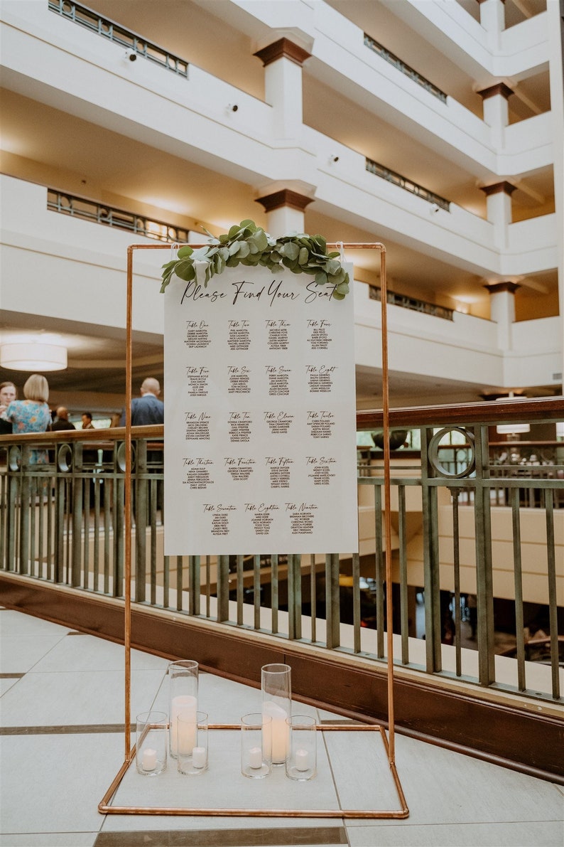 May include: A white seating chart with the text "Please Find Your Seat" at the top. The chart is displayed on a copper stand with greenery at the top. The chart lists table numbers and names of guests.