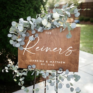 May include: A wooden sign with the text "The Reiners, Sabrina & Matthew Est. 2022" in white lettering. The sign is decorated with eucalyptus greenery and white flowers.