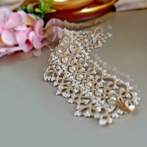 Pu&ograve; includere: Un delicato girocollo in pizzo dorato e bianco con un accento di cristallo.