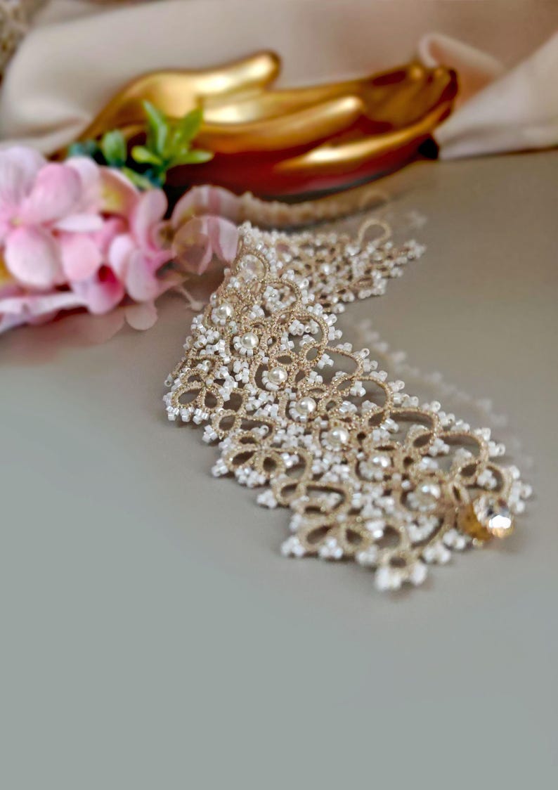 Pu&ograve; includere: Un delicato girocollo in pizzo dorato e bianco con un accento di cristallo.