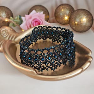 Pu&ograve; includere: Un girocollo in pizzo nero con perline blu iridescenti. Il girocollo &egrave; su una superficie dorata.