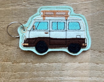 May include: A teal, white, and brown camper van keychain. The van has black wheels, a brown lower section, and a white upper section with teal windows. The keychain has a metal ring attached to the top left corner.