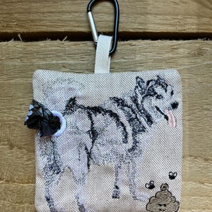 Könnte beinhalten: Beiger Hundekotbeutelspender mit schwarzem Karabinerhaken. Der Spender zeigt ein gesticktes Husky-Design mit schwarzen und grauen Details, eine herausgestreckte Zunge und ein Kack-Emoji mit Fliegen.