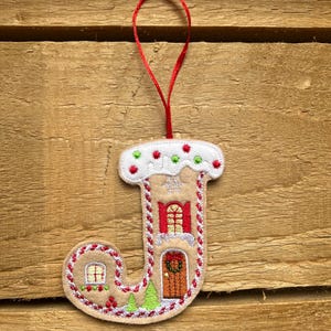 Op de afbeelding: Een beige vilt ornament in de vorm van de letter J, ontworpen als een peperkoekhuisje met witte glazuurranden, rood-witte zuurstokstrepen, een bruine deur met krans, rode luiken, een klein raam en groene dennenbomen met een rood lint.