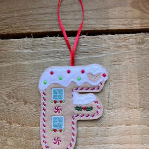 May include: A festive felt Christmas ornament in the shape of the letter "F", designed as a gingerbread house. It features white icing, red and green details, and a red ribbon. The background is a wooden surface.