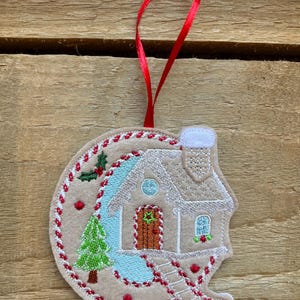 Può includere: Un ornamento circolare in feltro con ricamata una casa di pan di zenzero dotata di tetto bianco, porta marrone e camino. Il design include un pino verde, bacche rosse e uno sfondo azzurro, il tutto completato da un nastro rosso per appenderlo.