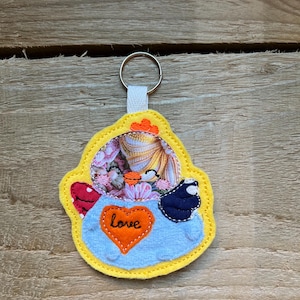 May include: A handmade keychain with a yellow border and a blue center. The keychain features a heart with the word "love" and a floral design. The keychain is attached to a silver metal ring with a white fabric loop.