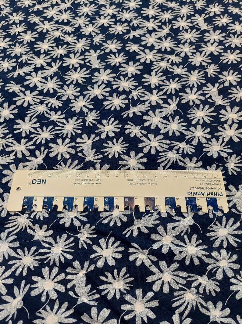 May include: Navy blue fabric with a white daisy print. The fabric is measured with a ruler that reads "Pifferi Anelio Schneidereibedarf Fermewaldstr 78 46145 Oberhausen Telefon 0208)437400 084374040 Telefax 208 Intermec www.pfende Mer infogasteri de"