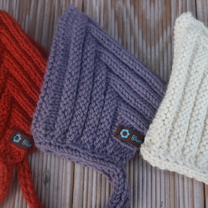 Könnte beinhalten: Drei gestrickte Baby-Mützen in Orange, Lila und Weiß mit einem Chevron-Muster. Jede Mütze hat ein kleines Etikett mit dem Wort "Blum" darauf.