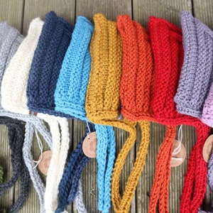 May include: A row of ten knitted hats in various colors, including gray, white, navy blue, light blue, mustard yellow, orange, red, purple, and pink. Each hat has a long knitted tie.