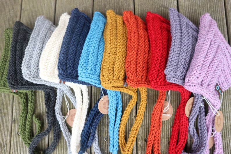 May include: A row of ten knitted hats in various colors, including gray, white, navy blue, light blue, mustard yellow, orange, red, purple, and pink. Each hat has a long knitted tie.