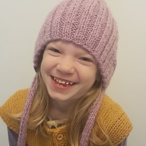 May include: A young child wearing a purple knitted hat with long ties, a yellow knitted vest, and a purple shirt. The child is smiling.