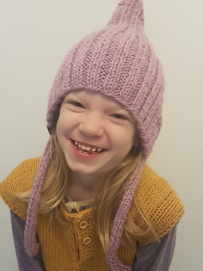 May include: A young child wearing a purple knitted hat with long ties, a yellow knitted vest, and a purple shirt. The child is smiling.