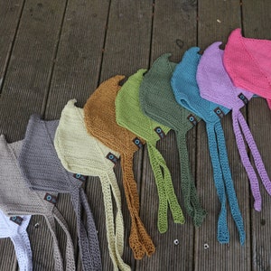 Puede incluir: Una colección de gorros tejidos a mano en varios colores, incluyendo blanco, gris, marrón, verde, azul, morado y rosa. Cada gorro tiene lazos largos y una pequeña etiqueta. Los gorros están colocados sobre una superficie de madera.