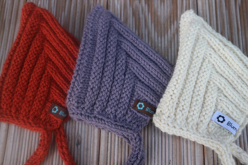 May include: Three knitted baby bonnets in orange, purple, and white with a chevron pattern. Each bonnet has a small tag with the word "Blum" on it.