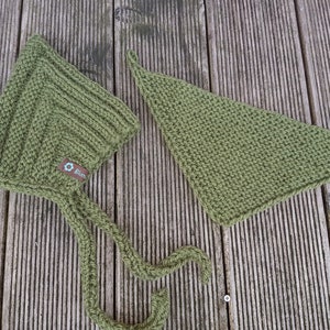 May include: A green knitted baby bonnet with a long tie and a matching green knitted triangle bib with a wooden button. Both items have a small brown label with a blue flower logo and the word "Bl&uuml;m" on it.