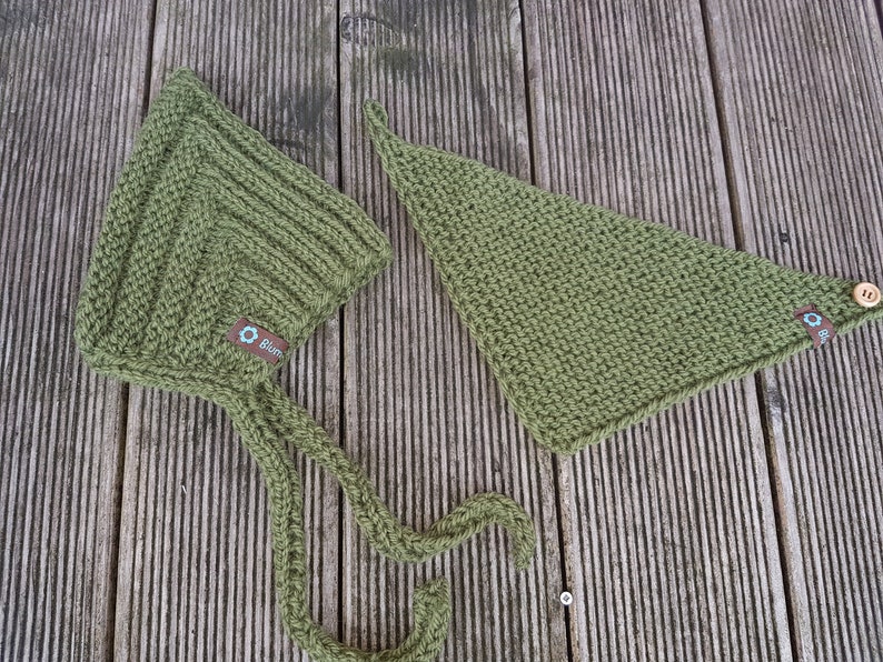 May include: A green knitted baby bonnet with a long tie and a matching green knitted triangle bib with a wooden button. Both items have a small brown label with a blue flower logo and the word "Bl&uuml;m" on it.