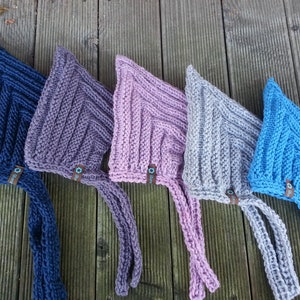 May include: Five knitted baby bonnets in different colors: navy blue, purple, pink, gray, and light blue. Each bonnet has a long tie and a small tag with the word "Bloom" on it.