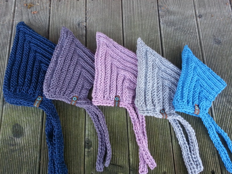 May include: Five knitted baby bonnets in different colors: navy blue, purple, pink, gray, and light blue. Each bonnet has a long tie and a small tag with the word "Bloom" on it.