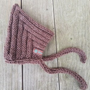 May include: A brown knitted baby bonnet with ties. The bonnet has a small tag with the word "Blomme" on it.