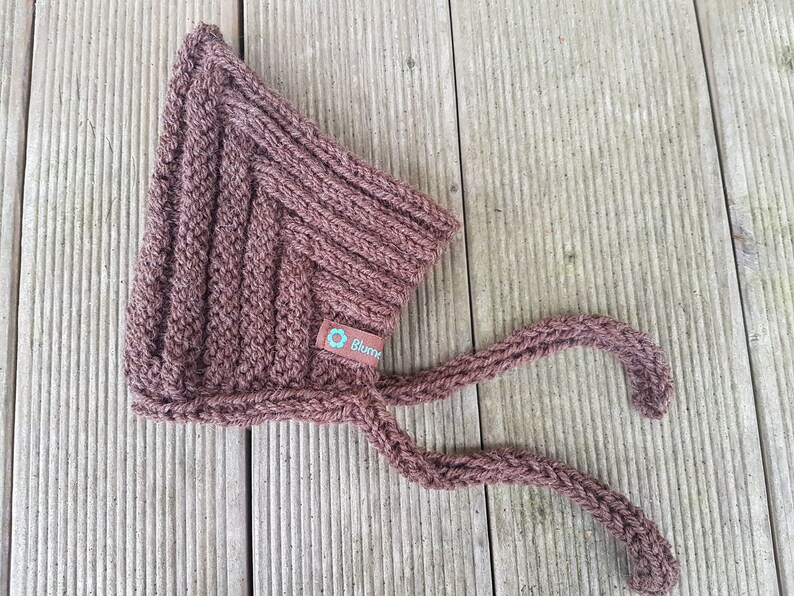 May include: A brown knitted baby bonnet with ties. The bonnet has a small tag with the word "Blomme" on it.