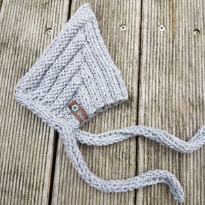 May include: Gray knitted baby bonnet with a long tie. The bonnet has a small label with the word "Bl&uuml;mchen" on it.