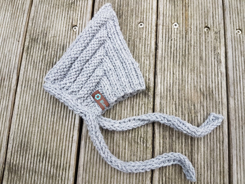 May include: Gray knitted baby bonnet with a long tie. The bonnet has a small label with the word "Bl&uuml;mchen" on it.