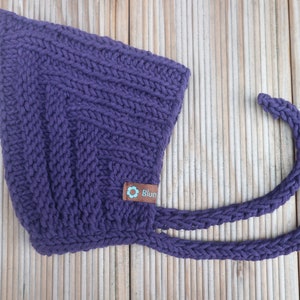 Peut inclure: Bonnet en tricot pour bébé de couleur violette avec une attache. Le bonnet a une petite étiquette marron avec une fleur et le mot "Blum" dessus.