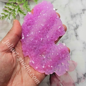 May include: A handcrafted, irregular-shaped, light purple crystal-like object. The object is held in a hand and has a textured surface with many small, reflective facets. The background includes greenery and the text "juliart-studio.com".