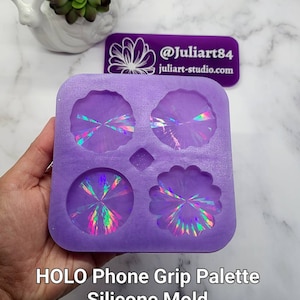 May include: Purple silicone mold with four different shapes for making phone grips. The shapes include a circle, a flower, and two different diamond shapes. The mold is filled with iridescent resin, showing off the holographic effect.