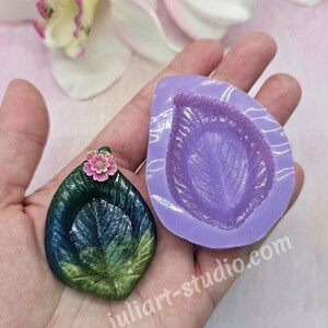 Peut inclure: Un pendentif artisanal en forme de feuille avec un petit accent de fleur rose, à côté d'un moule en silicone violet. Le pendentif est vert et bleu avec des reflets dorés. L'adresse du site Web juliart-studio.com est visible.