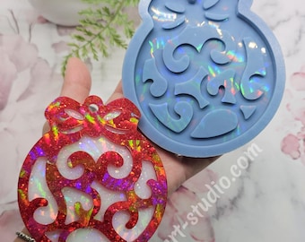 May include: A blue silicone mold and a red glitter filled resin ornament, both shaped like a Christmas ornament with a bow on top. The ornament has a swirl design.