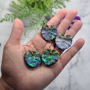 May include: A pair of iridescent green and purple leaf-shaped earrings with gold hooks. The earrings are made of resin and have a textured surface.
