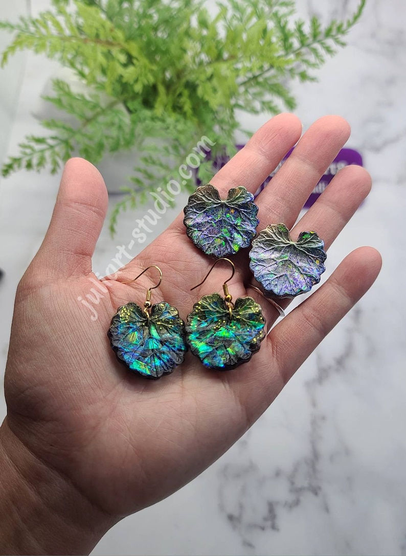 May include: A pair of iridescent green and purple leaf-shaped earrings with gold hooks. The earrings are made of resin and have a textured surface.