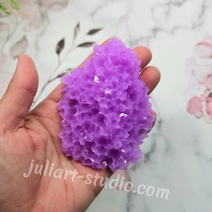 May include: A vibrant purple crystal cluster held in a hand. The crystal has a textured, irregular surface with small, reflective facets. The website julia-rt-studio.com is visible in the image.