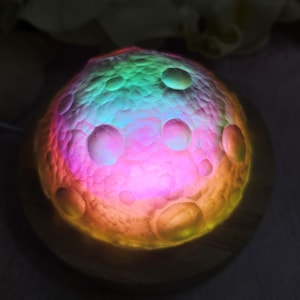 May include: A spherical moon-shaped lamp featuring a textured surface with craters, illuminated by a vibrant, multi-colored LED light that transitions through shades of pink, blue, green, and yellow, resting on a circular wooden base.